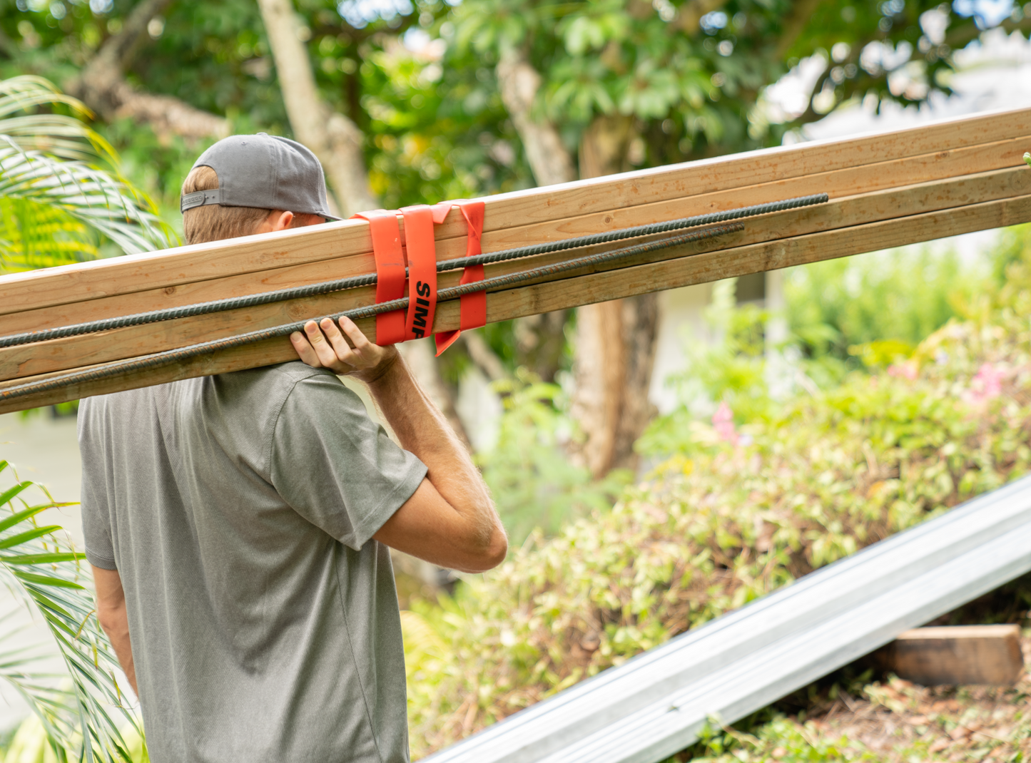Person bærer treverk og armeringsjern buntet med røde Simple Strap heavy duty stropper utendørs.